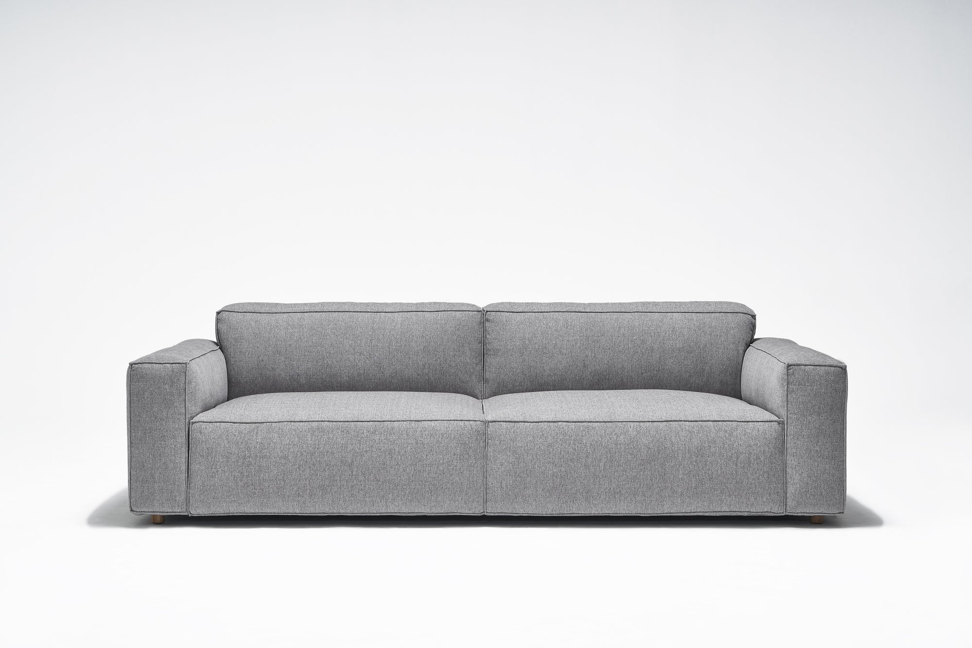 The Baker Sofa | Fabric by Wooden Stories features a contemporary gray design with wide armrests, a low backrest, and plush feather filling, displayed against a plain white background.