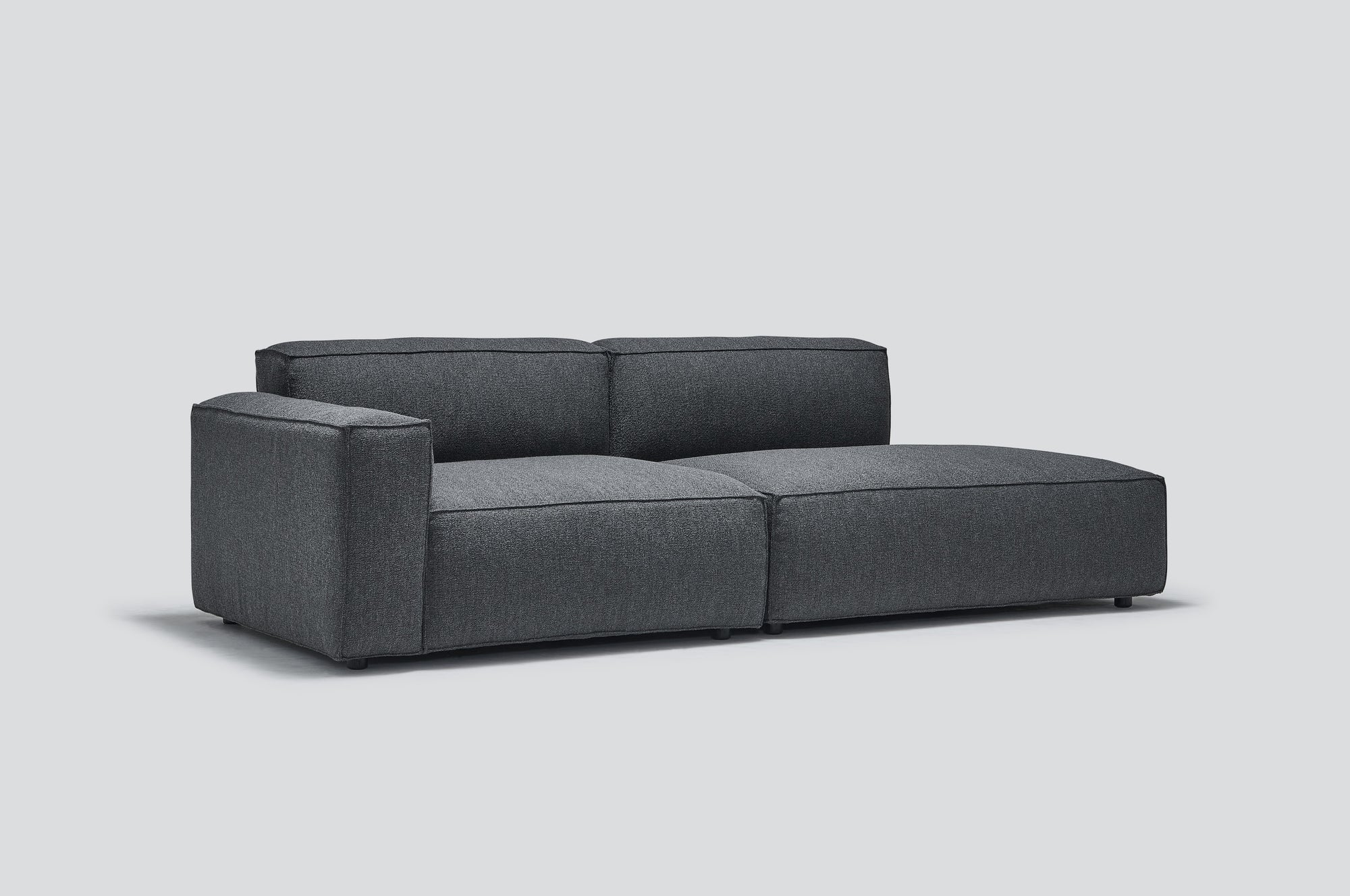 The Baker Modular Sofa | Fabric • M1 by Wooden Stories features a modern dark gray design with a low backrest and wide seat cushions, showcased against a plain light gray background.