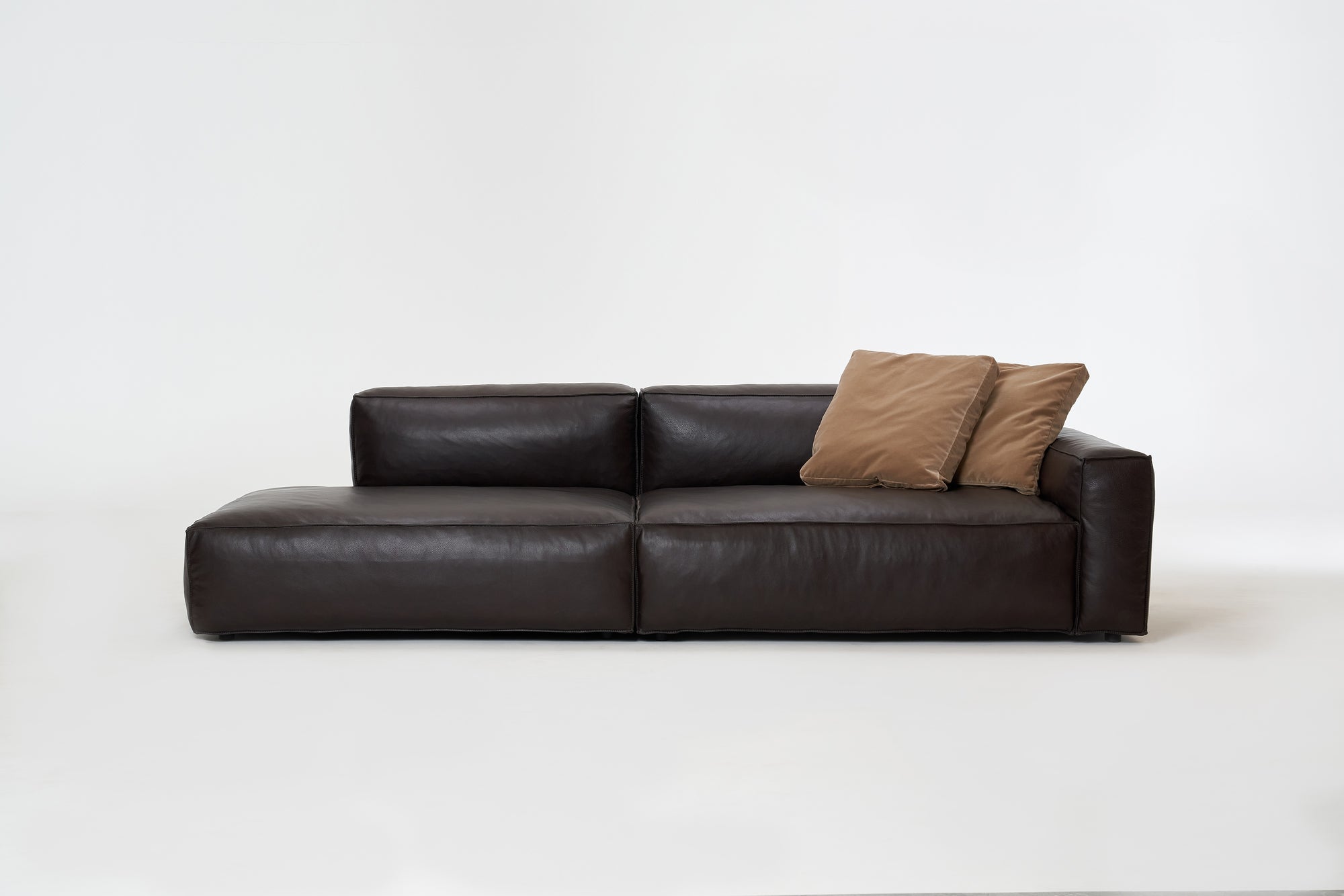 The Baker Modular Sofa | Leather • M2 by Wooden Stories is a modern dark brown leather sofa with a low backrest, left-side chaise, and two light brown throw pillows, shown against a plain white background.