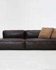 The Baker Modular Sofa | Leather • M2 by Wooden Stories is a modern dark brown leather sofa with a low backrest, left-side chaise, and two light brown throw pillows, shown against a plain white background.