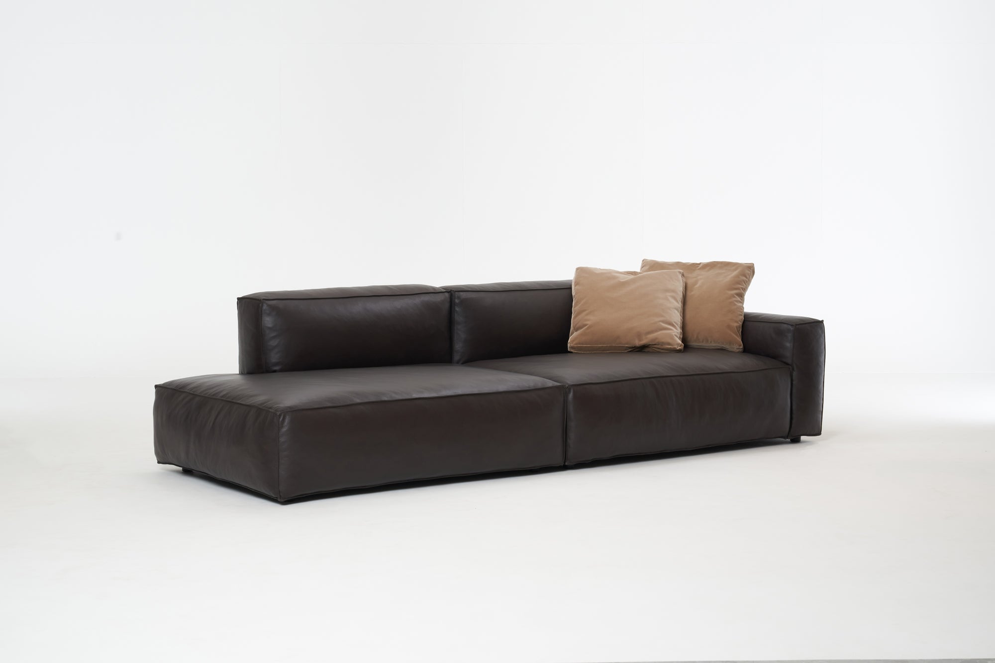 The Baker Modular Sofa | Leather • M2 by Wooden Stories is a modern dark brown leather sofa with two light brown throw pillows, showcased against a plain white background.