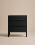 The Nell Tall Dresser by Wooden Stories, part of the Danish Collection, is a modern black wooden nightstand featuring three drawers with clean lines, premium metal runners, and a handleless design. Shown against a plain beige background, it offers stylish yet practical storage for your bedroom.