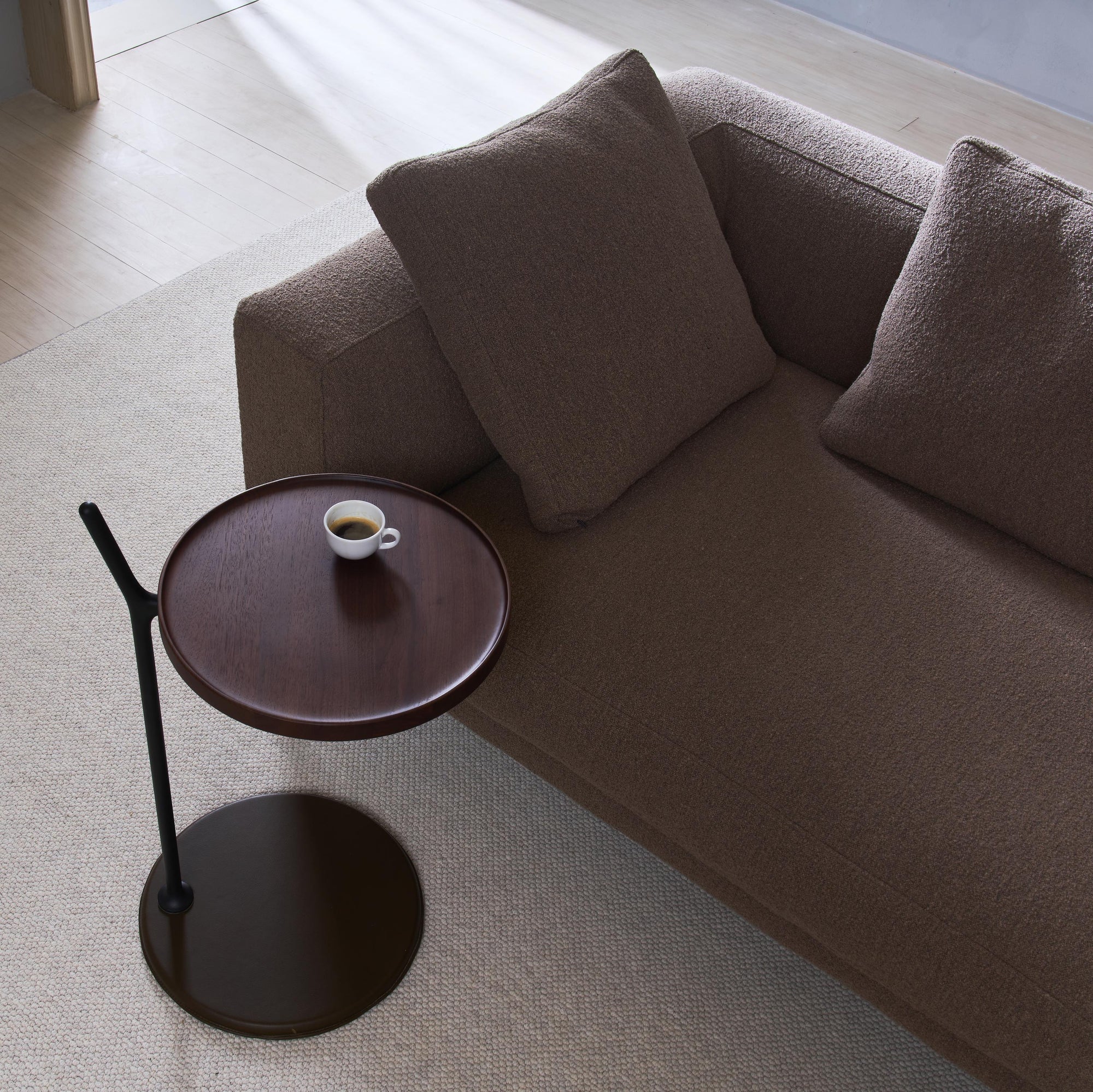 A brown sofa with two cushions sits beside the Wooden Stories Bystander Side Table, which holds a small cup of espresso. The scene features a light rug and wooden flooring in a minimalist room.