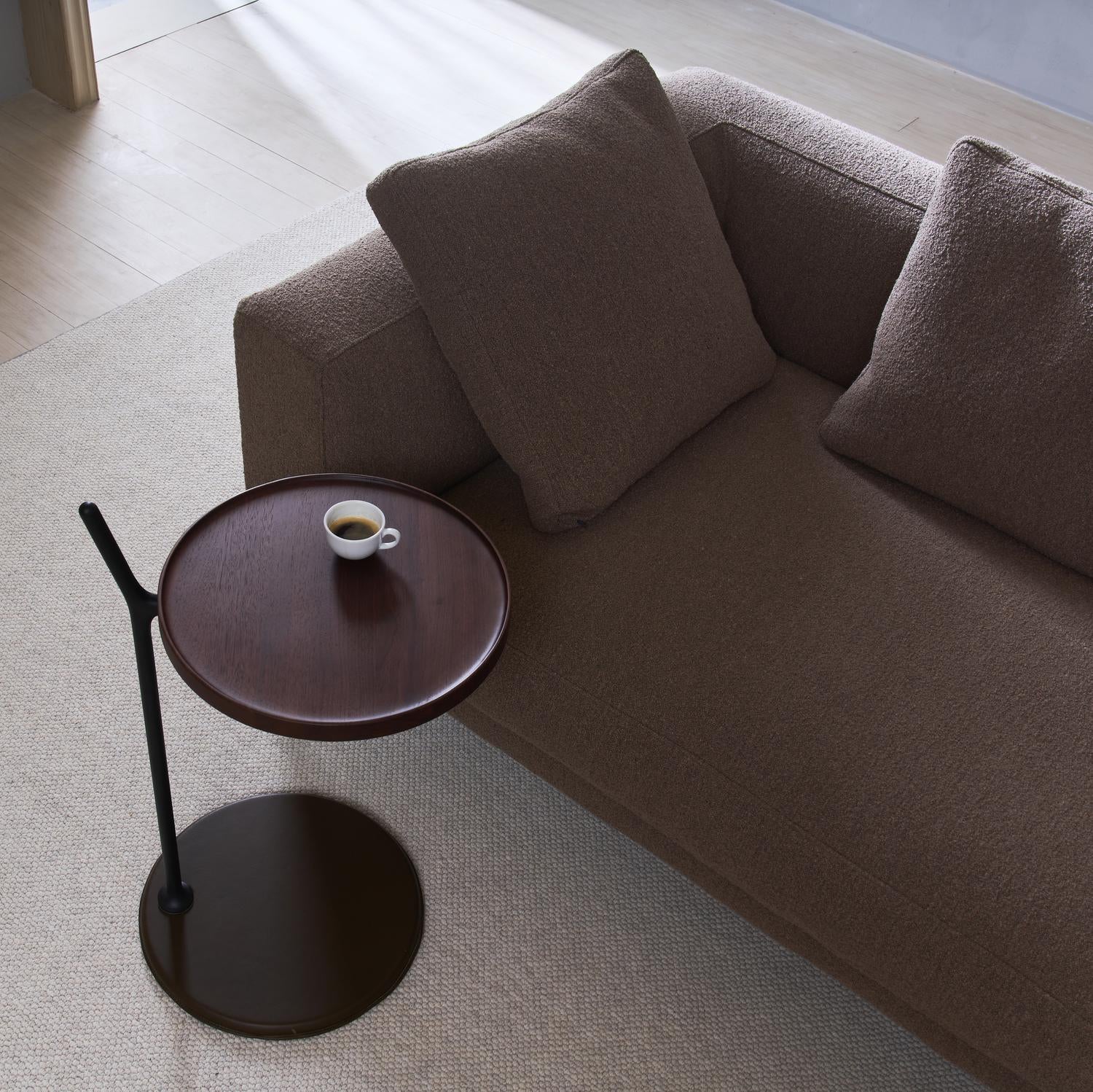 A brown sofa with two cushions sits next to the Wooden Stories Bystander Side Table—a small, solid wood piece from the Danish Collection that holds a cup of espresso. Sunlight shines on the light wood floor and beige rug.