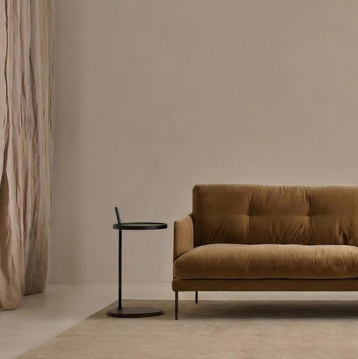 A minimalist living room featuring a brown velvet sofa, the round Bystander Side Table by Wooden Stories, beige curtains, and a neutral area rug atop a light floor. The space exudes calm with muted tones and elegantly simple furnishings.