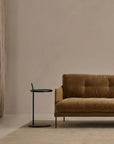 A minimalist living room featuring a brown velvet sofa, the round Bystander Side Table by Wooden Stories, beige curtains, and a neutral area rug atop a light floor. The space exudes calm with muted tones and elegantly simple furnishings.