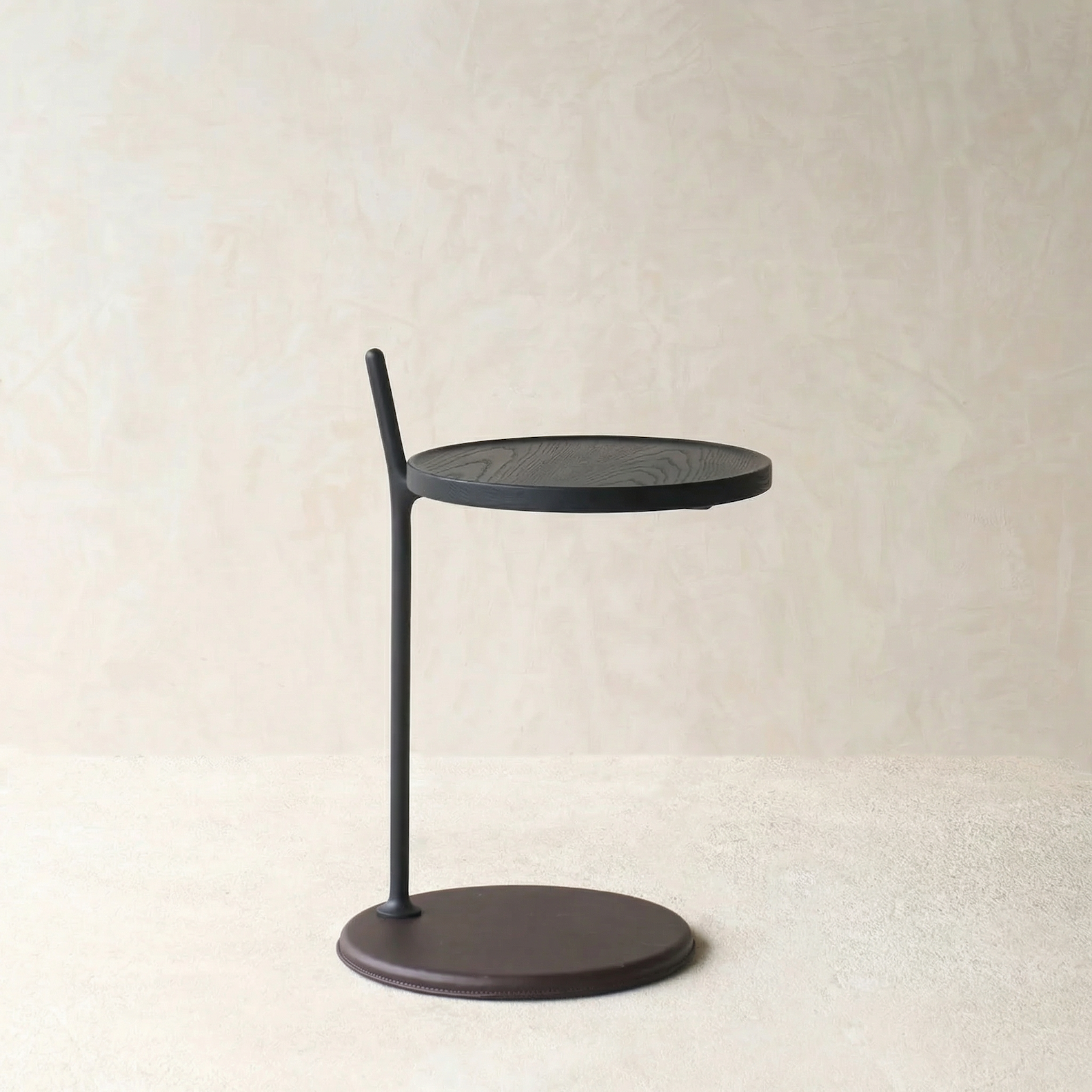 The Bystander Side Table by Wooden Stories features a round solid wood top and base with a single black metal rod, offering modern minimalist style against a neutral backdrop.