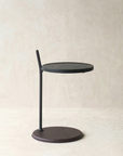The Bystander Side Table by Wooden Stories features a round solid wood top and base with a single black metal rod, offering modern minimalist style against a neutral backdrop.