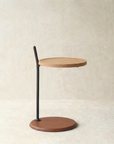 The Wooden Stories Bystander Side Table features a modern, minimalist design with a solid wood top, slim black metal support, and circular brown base from the Danish Collection, set against a neutral textured background.