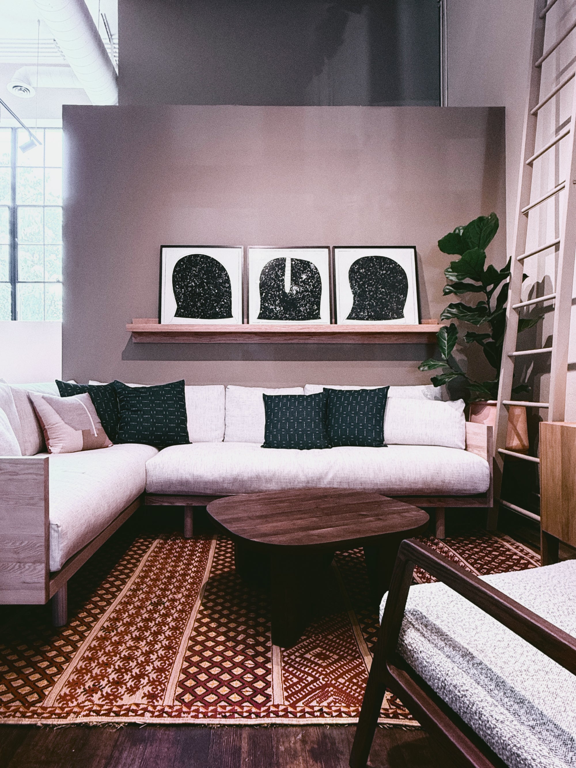 A modern living room with a light sectional sofa, patterned cushions, a round wooden table, a patterned rug, and "Celestial Reflections" by ROBERT BRADY framed on the shelf above the couch. A plant and ladder shelf sit in the corner.