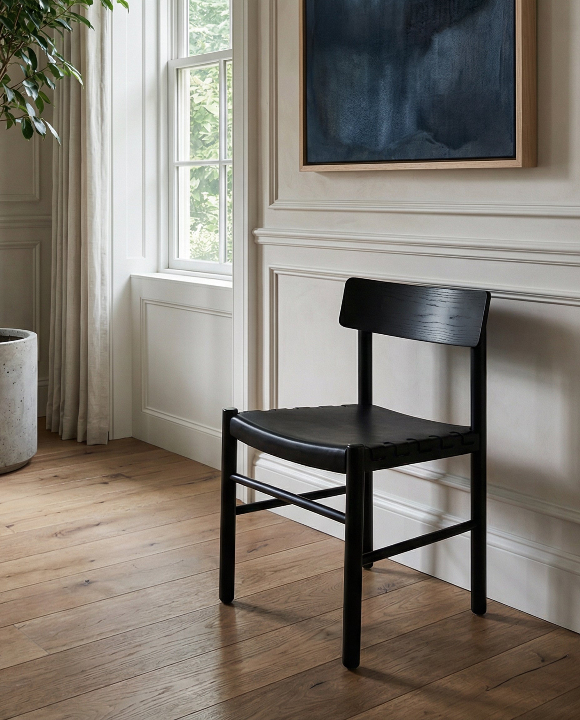 A SOBU Cesco Dining Chair in Black Ash is placed on a light wooden floor near a large window with white curtains. A potted plant sits nearby and a dark abstract painting hangs on the white paneled wall behind the chair.