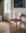 A Cesco Dining Chair by SOBU, crafted from solid white ash and featuring a heritage red top-grain leather seat, sits on a wooden floor near a large window, accompanied by a potted plant and an abstract blue painting on the wall behind it.