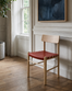 A Cesco Dining Chair by SOBU, crafted from solid white ash and featuring a heritage red top-grain leather seat, sits on a wooden floor near a large window, accompanied by a potted plant and an abstract blue painting on the wall behind it.
