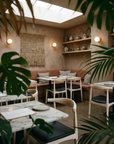 A cozy, modern cafe with marble tables, SOBU Copenhagen Dining Chairs in White Ash, and a leather seat bench along the wall. Potted plants, ceramic vases on shelves, and a woven wall hanging create a warm, inviting atmosphere.