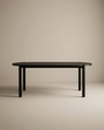 The Cove Dining Table | Black Oak by Wooden Stories, featuring a minimalist design with rounded corners and four cylindrical legs in solid American oak, stands in an empty room with plain beige walls and floor.