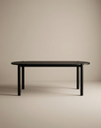 The Cove Dining Table | Black Oak by Wooden Stories, featuring a minimalist design with rounded corners and four cylindrical legs in solid American oak, stands in an empty room with plain beige walls and floor.