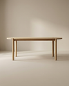 The Wooden Stories Cove Dining Table | Light Oak features solid American oak with rounded edges and four cylindrical legs, shown against a neutral beige background in a softly lit room.