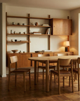 A modern dining room features Wooden Stories’ Poise Dining Chair in Light Oak with Leather, a solid oak table and sideboard, wall shelves with ceramics and books, all set against a light wall and parquet floor.