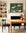 A modern dining room with a wooden table, SOBU’s Copenhagen Dining Chairs in Black Ash with leather seats, an orange vase with foliage, green art above a white fireplace, shelves of pottery and framed art, and two orange candles on the mantel.