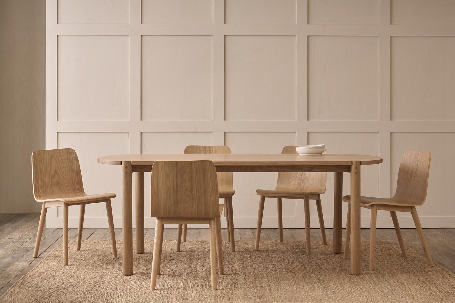 A minimalist dining room features the Wooden Stories Cove Dining Table in Light Oak, five matching wooden chairs, and a simple bowl, set against a beige panel wall with a neutral woven rug on the floor.