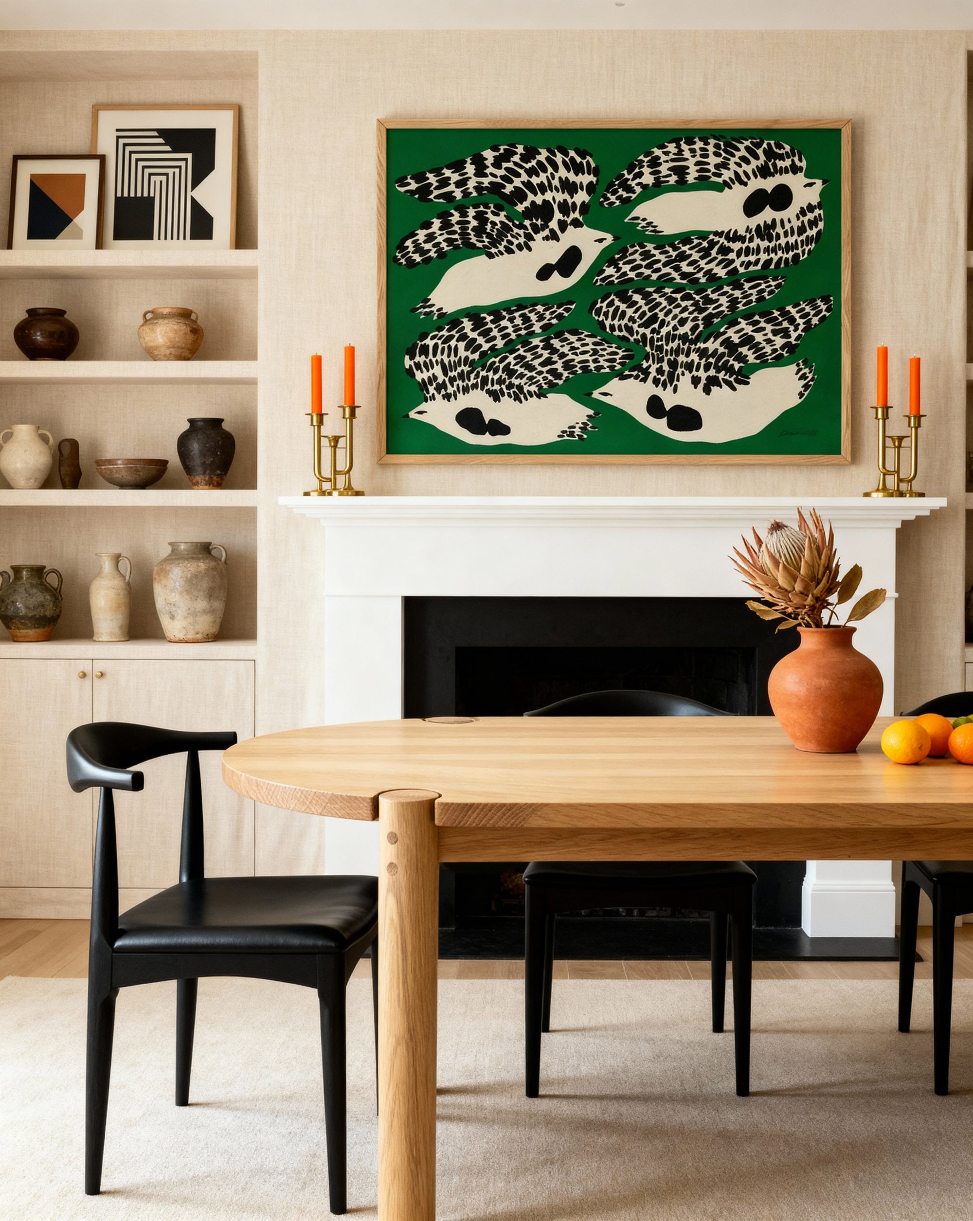 A modern dining room features the Wooden Stories Cove Dining Table in light oak, black chairs, a vase with dried flowers and oranges, pottery on shelves, and bold bird art above a white fireplace set against a green background.