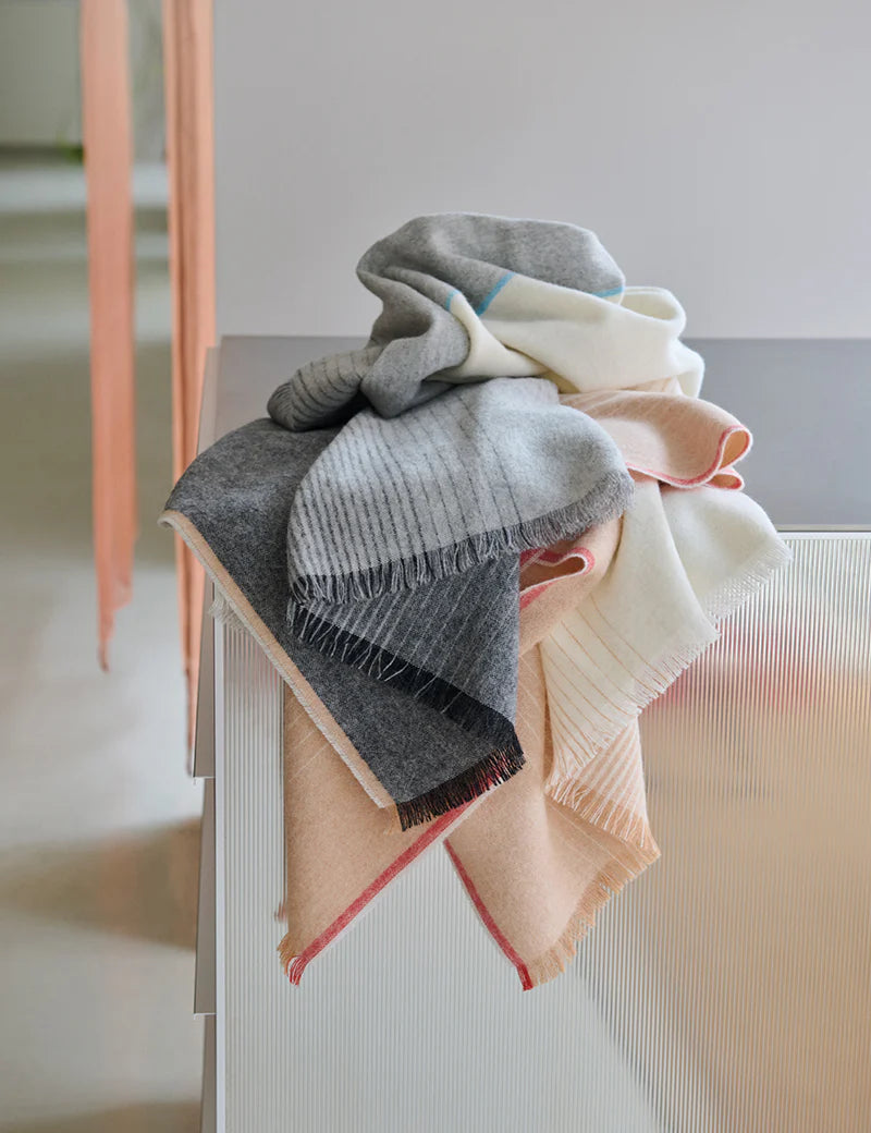 The Carte Postale Throw - Grey by Elvang Denmark, crafted from recycled wool in soft gray tones, is neatly folded and draped over the corner of a textured white cabinet, adding cozy elegance to a modern, minimal space.