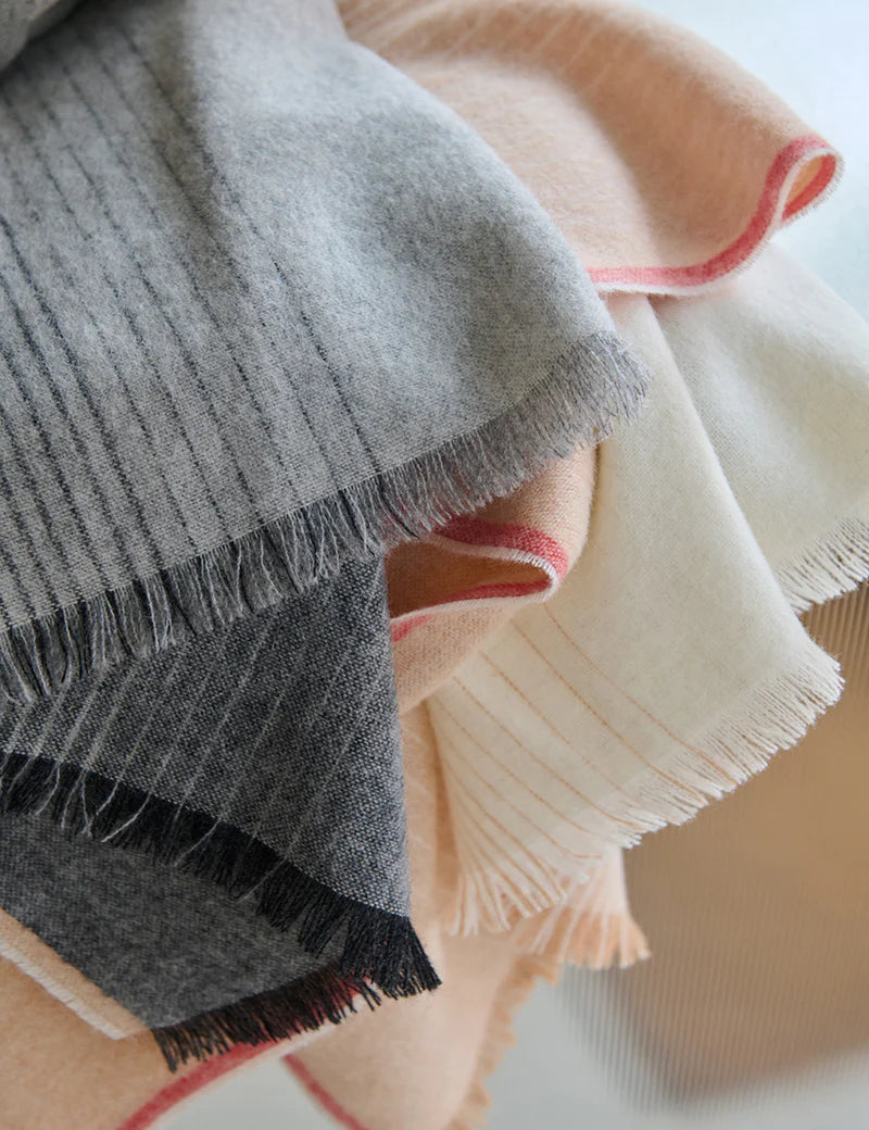 A close-up of the Elvang Denmark Carte Postale Throw - Grey, crafted from recycled wool in soft gray with subtle pinstripes and fringed edges. The fabric looks invitingly soft and cozy, accented with hints of cream, peach, and red.