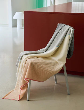 The Carte Postale Throw - Grey by Elvang Denmark, featuring soft recycled wool in subtle tones, is draped over a white chair with a modern red table and minimalistic decor in the background.