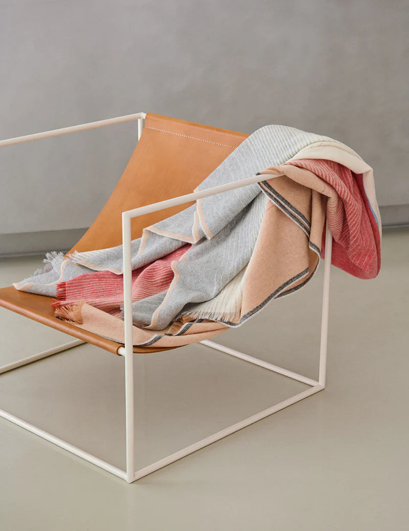A modern chair with a tan leather seat and white metal frame is draped with the Carte Postale Throw - Red Grey by Elvang Denmark, a soft alpaca wool blanket in gray, pink, and beige tones, set against a plain background.