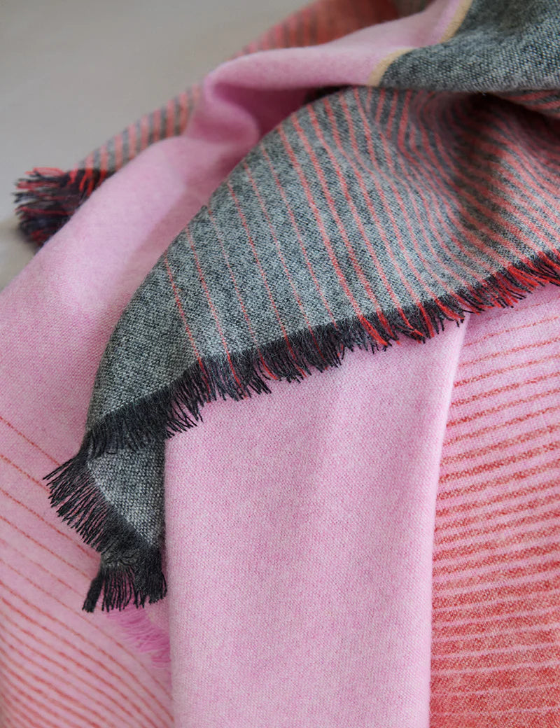 A close-up of the Elvang Denmark Carte Postale Throw - Red Pink shows overlapping sections: one in gray with red stripes and fringe, the other in light pink with subtle red stripes, highlighting its soft texture and cozy look.