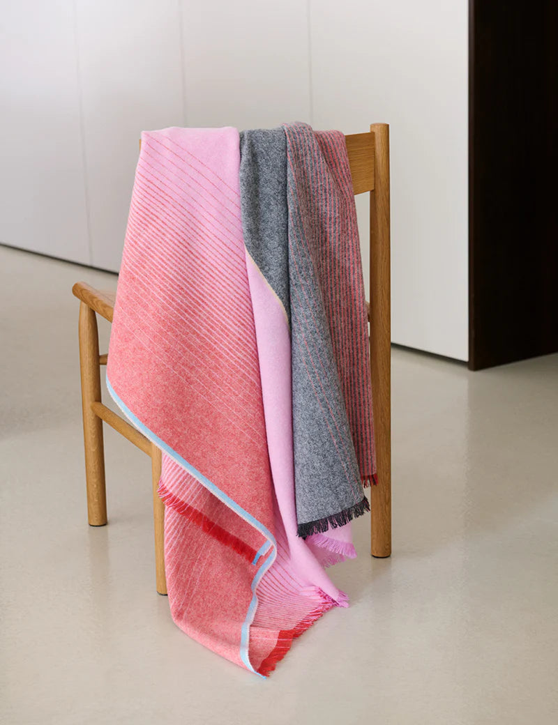 A wooden chair holds two blankets: the Elvang Denmark Carte Postale Throw in red and pink with blue trim, and a gray subtly patterned throw. The chair is on a light floor near white walls.