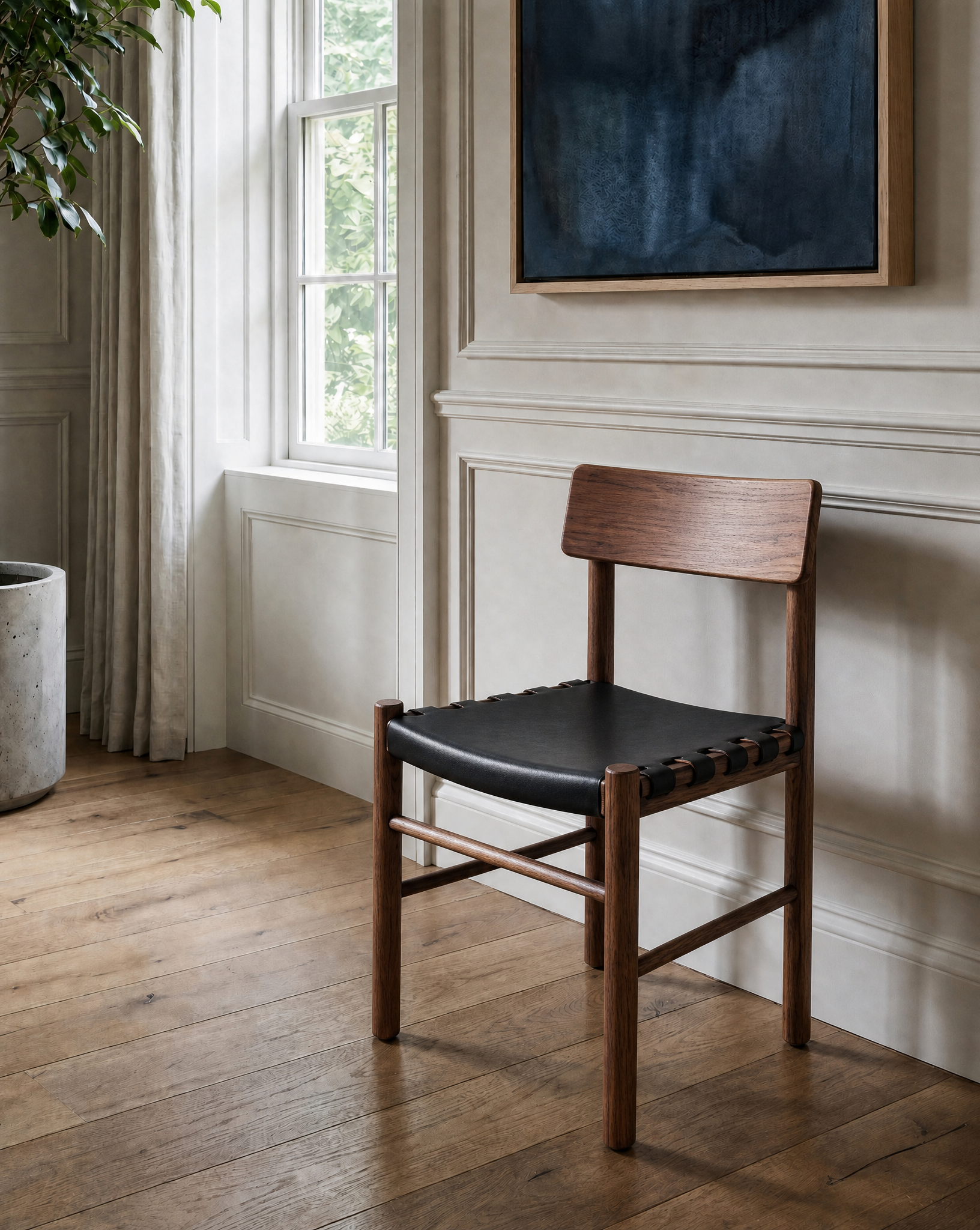 A SOBU Cesco Dining Chair in solid American black walnut with a top-grain leather seat sits on a hardwood floor beside a window, accompanied by a large potted plant and a blue abstract painting in a bright, minimal room with white paneled walls.
