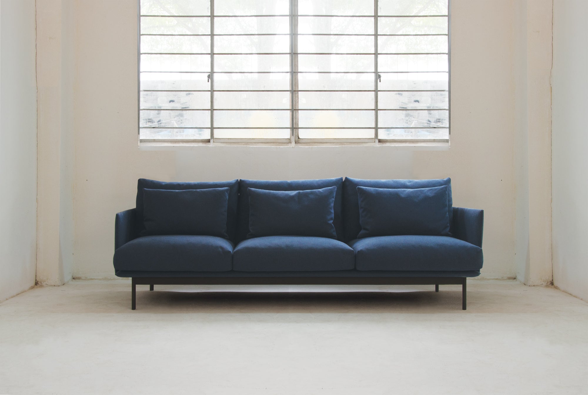A modern living space showcases the Cherry Sofa 95" | Fabric by Wooden Stories, with Danish lines and plush cushions, set against a white wall beneath a large window with metal bars in a bright, minimal room.