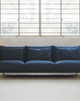 A modern living space showcases the Cherry Sofa 95" | Fabric by Wooden Stories, with Danish lines and plush cushions, set against a white wall beneath a large window with metal bars in a bright, minimal room.