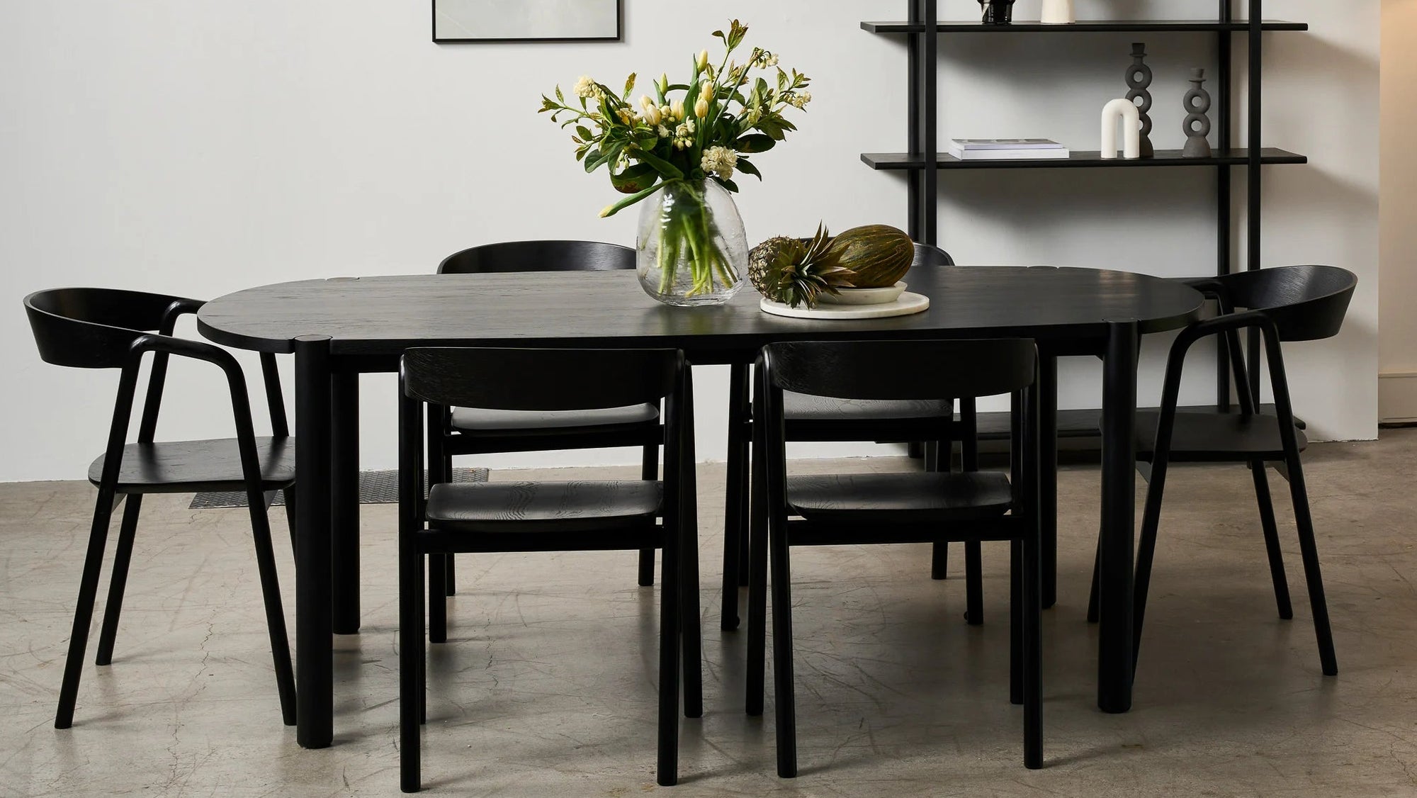 A modern dining room features the Wooden Stories Cove Dining Table in Black Oak, six black chairs, a flower vase, black shelves with decor, a wall mirror, a white armchair, a floor lamp, and minimalist art for an elevated dining experience.