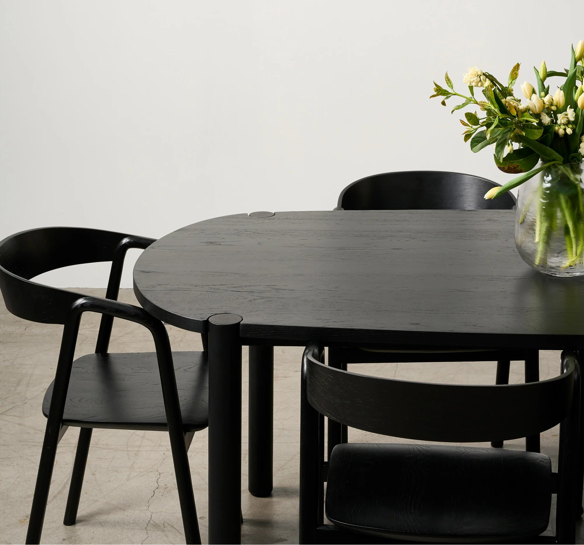 A minimalist dining area features the Wooden Stories Cove Dining Table in Black Oak, three black chairs, a glass vase with white and yellow flowers, and a framed hand photo on the white wall—perfect for an elevated dining experience.