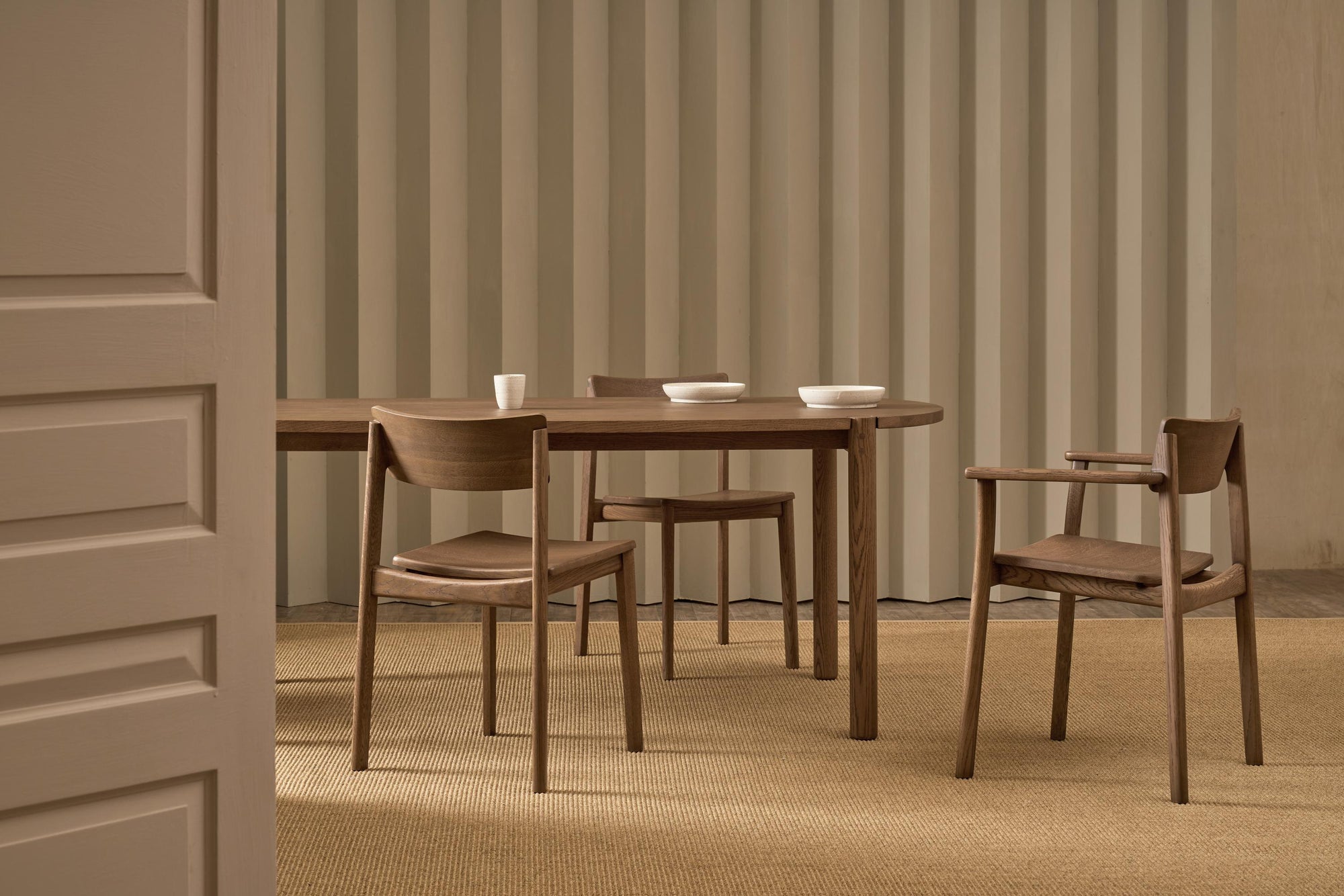 A minimalist dining room showcases the Cove Dining Table in Espresso Oak by Wooden Stories with three chairs on a tan rug. White dishes top the table, complementing a ribbed beige wall and open door for subtle Sydney Harbor vibes.