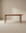 The Wooden Stories Cove Dining Table | Latte Oak features a minimalist design, meticulously crafted from solid American oak with rounded edges and four substantial cylindrical legs. Displayed against a neutral beige background, it is ideal for both residential spaces and contract-grade environments.