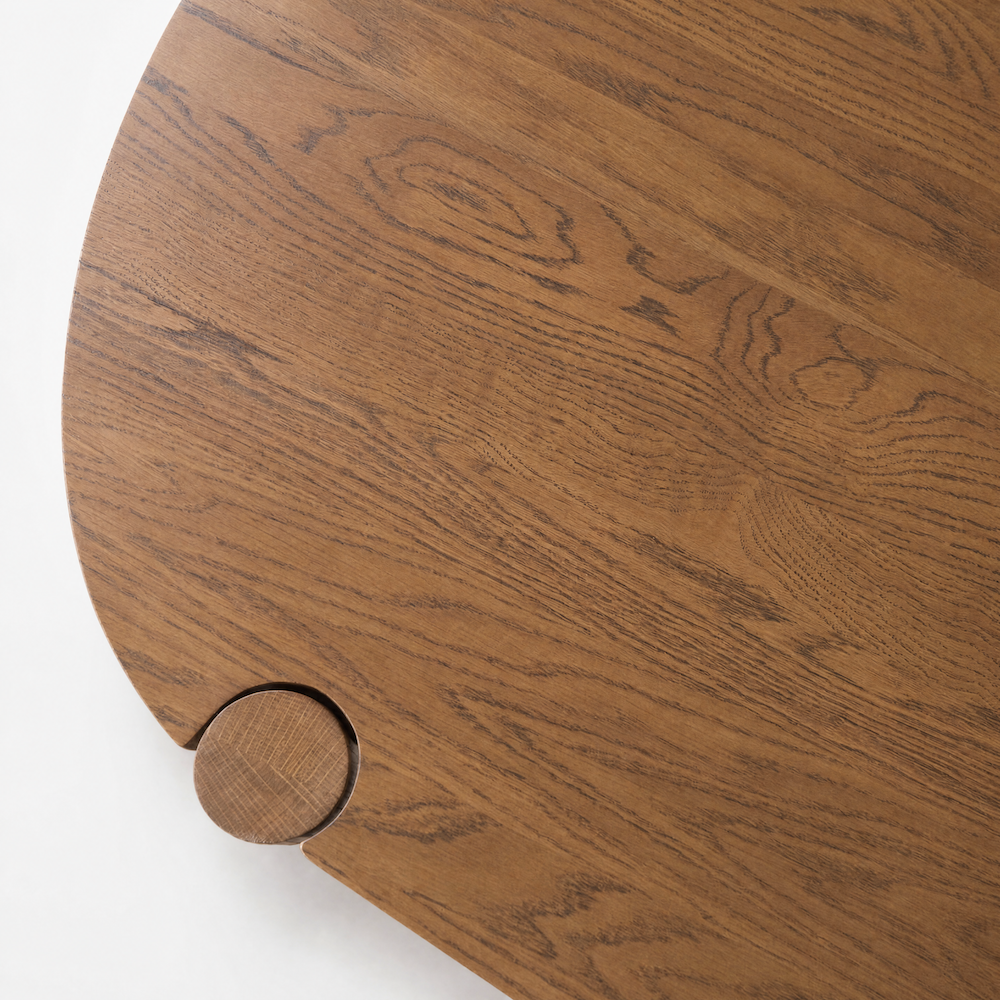 A close-up of a round wooden table crafted from solid American oak in the warm Latte Oak finish, with visible wood grain and a small circular protrusion on the edge—highlighting the contract-grade quality of the Cove Dining Table by Wooden Stories.