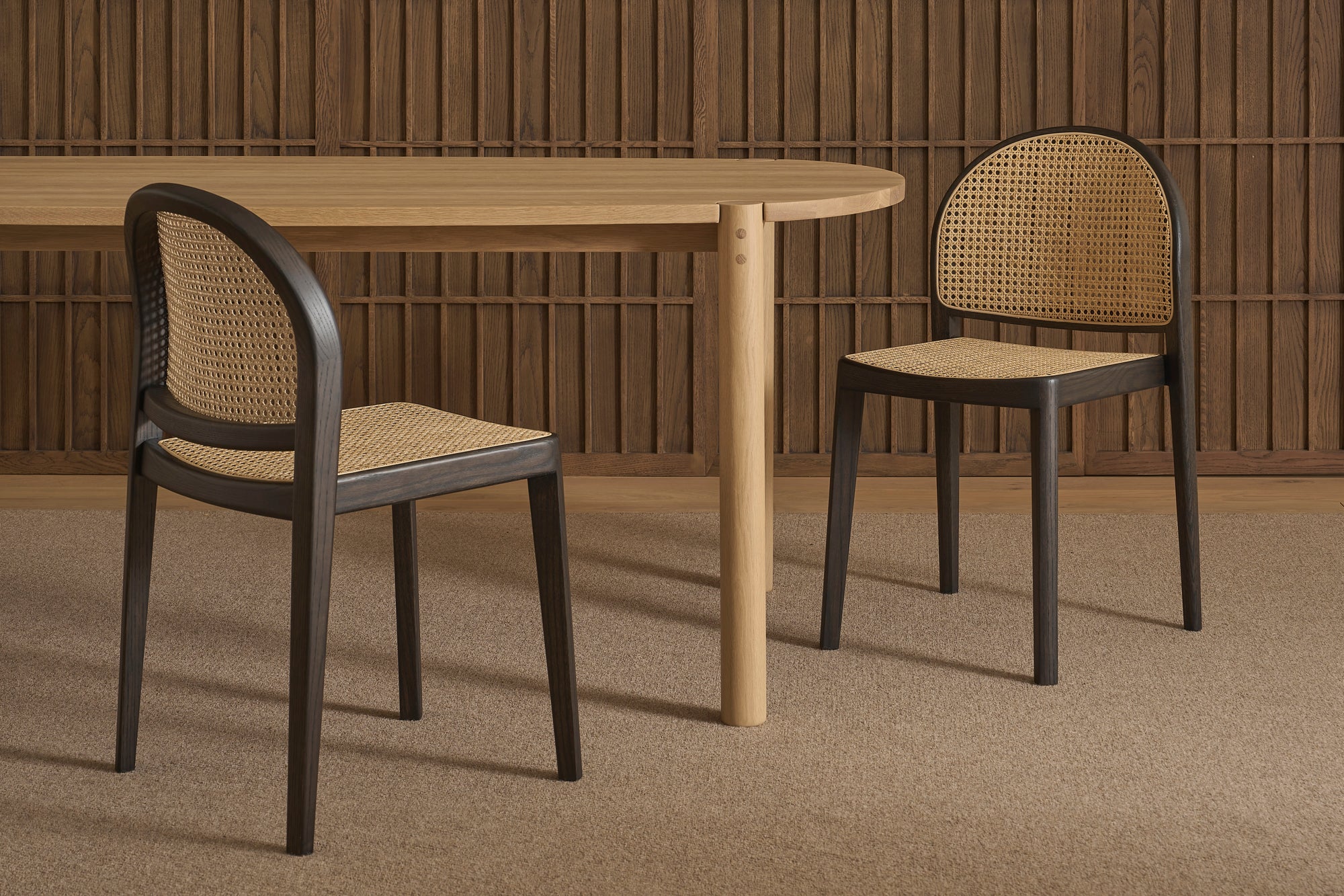 Two contract-grade Luk Dining Chairs by Wooden Stories, featuring cane weave rattan seats and backs, sit beside a light wooden table against vertical wood slats. The room is finished with a neutral carpeted floor.