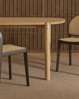 Two contract-grade Luk Dining Chairs by Wooden Stories, featuring cane weave rattan seats and backs, sit beside a light wooden table against vertical wood slats. The room is finished with a neutral carpeted floor.