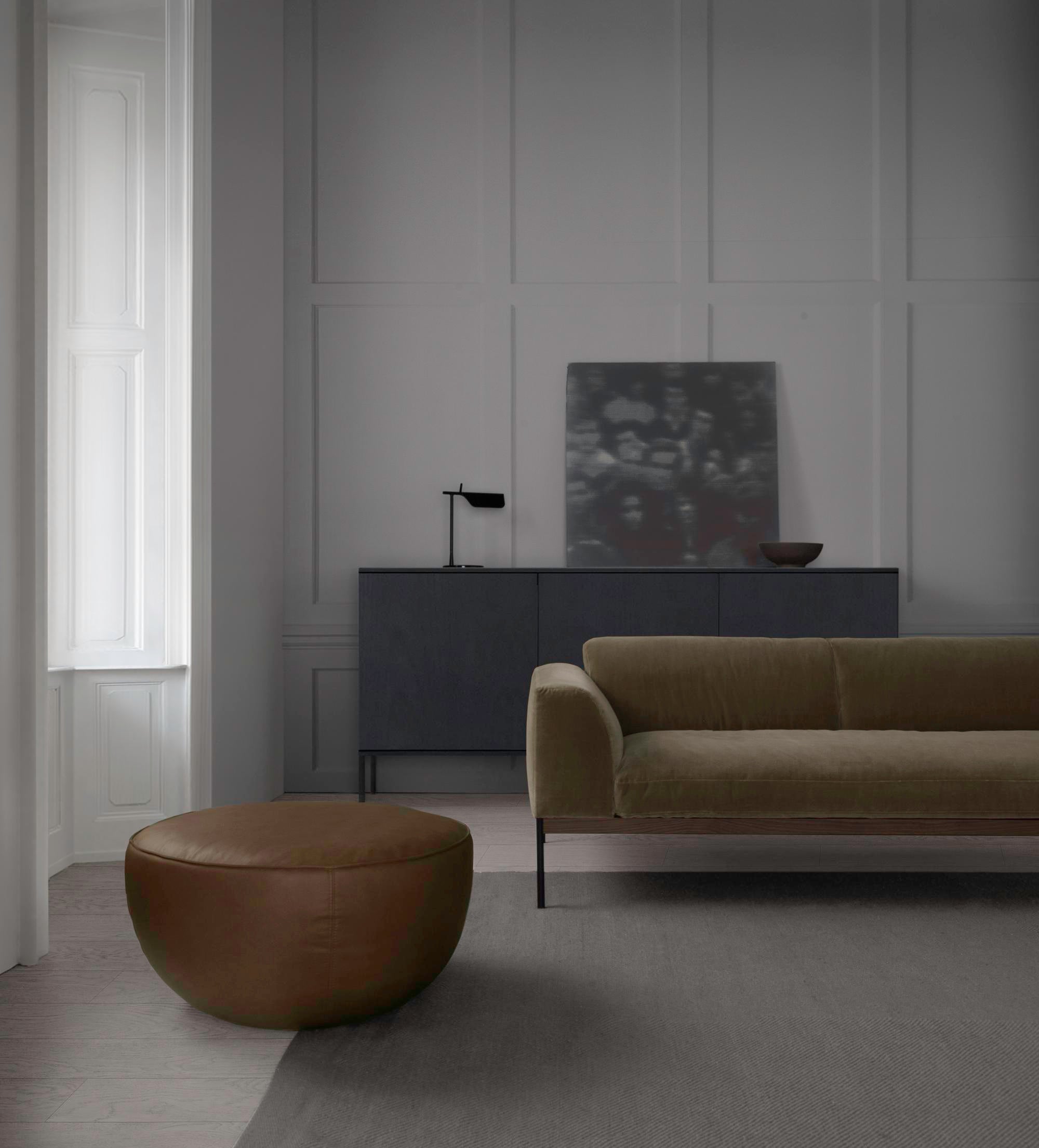 A minimalist living room features the Wooden Stories Department Sofa 93&quot; in fabric, a brown leather ottoman with solid wood base, dark sideboard, lamp, bowl, and abstract art against light gray paneled walls with natural light from a tall window.