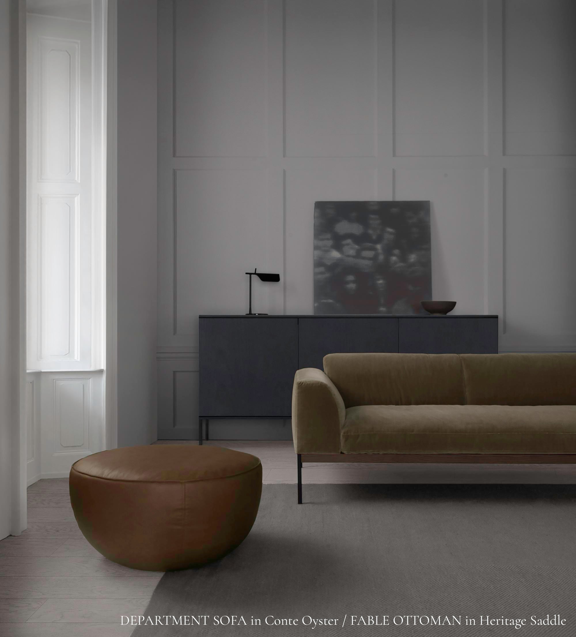A minimal living room features the Wooden Stories Department Modular Sofa | Fabric • M6 Left in beige, a round brown ottoman, dark sideboard, abstract art, table lamp, and bowl. Sunlight from tall windows highlights the neutral modern decor.
