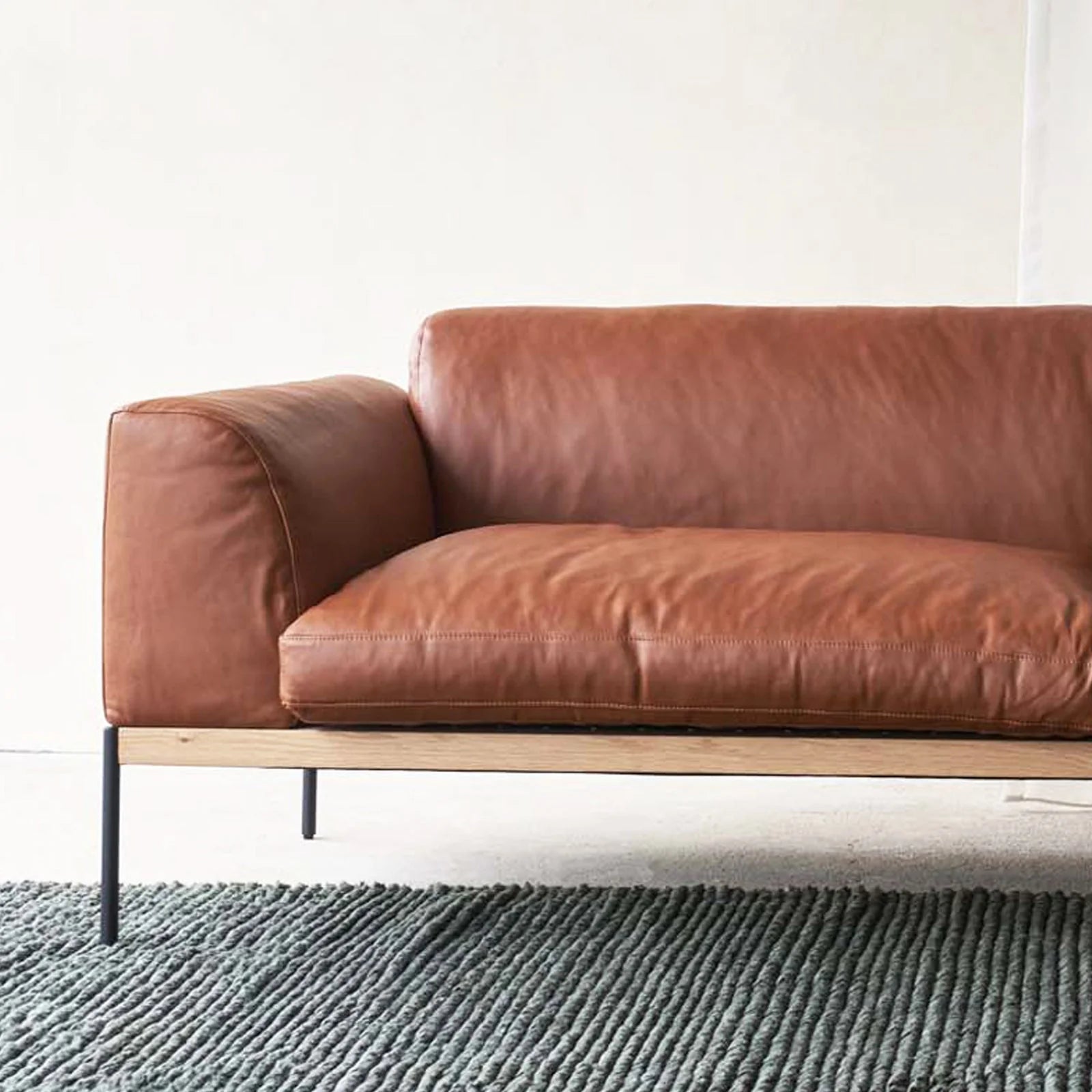 The Department Modular Sofa | Leather • M5 Right by Wooden Stories, with a solid wood base and black metal legs, sits stylishly on a textured gray rug against a plain light wall.