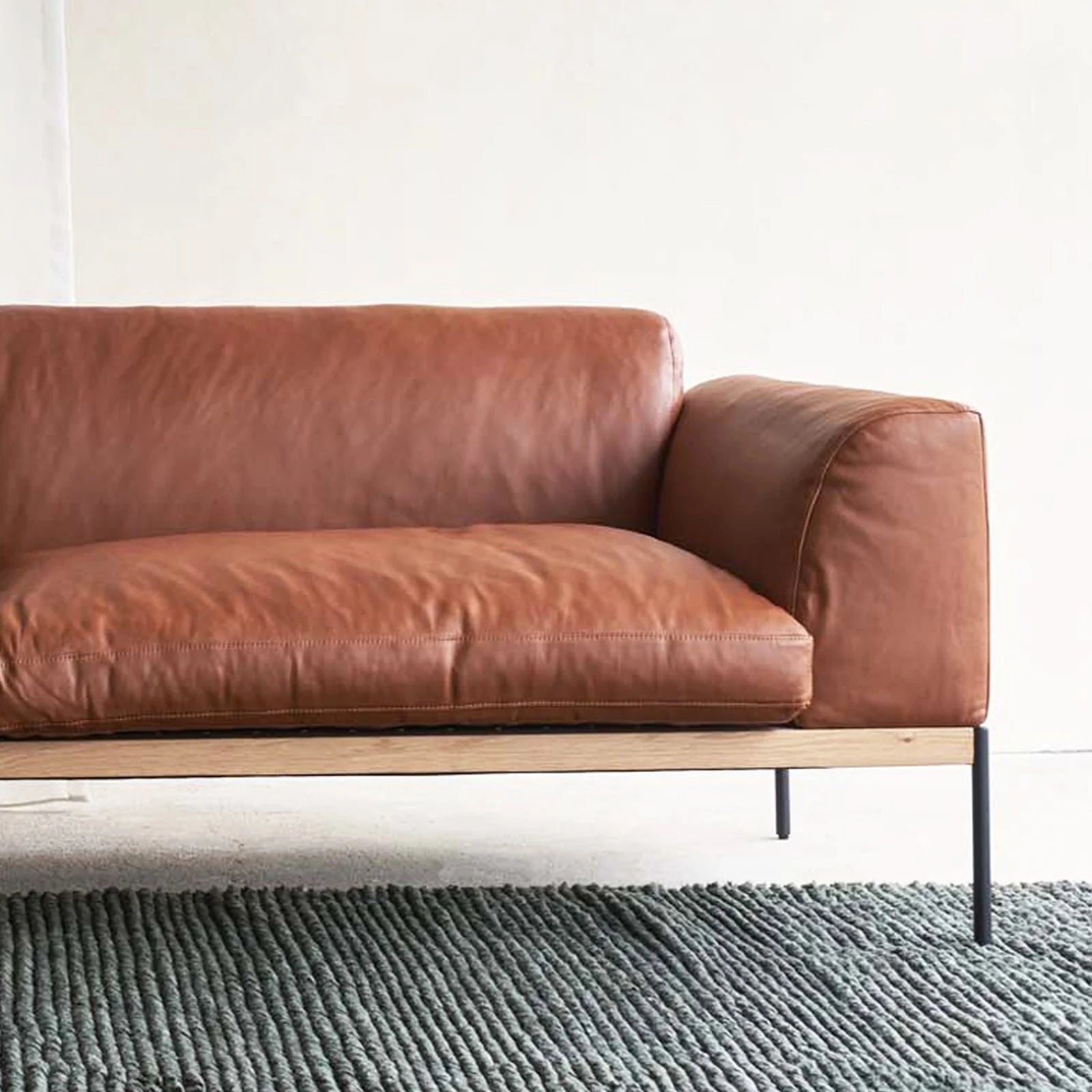 The Department Modular Sofa | Leather • M4 Left by Wooden Stories features a modern brown leather design with a solid wood base and thin black metal legs, positioned on a textured gray rug against a light wall, evoking Danish style.