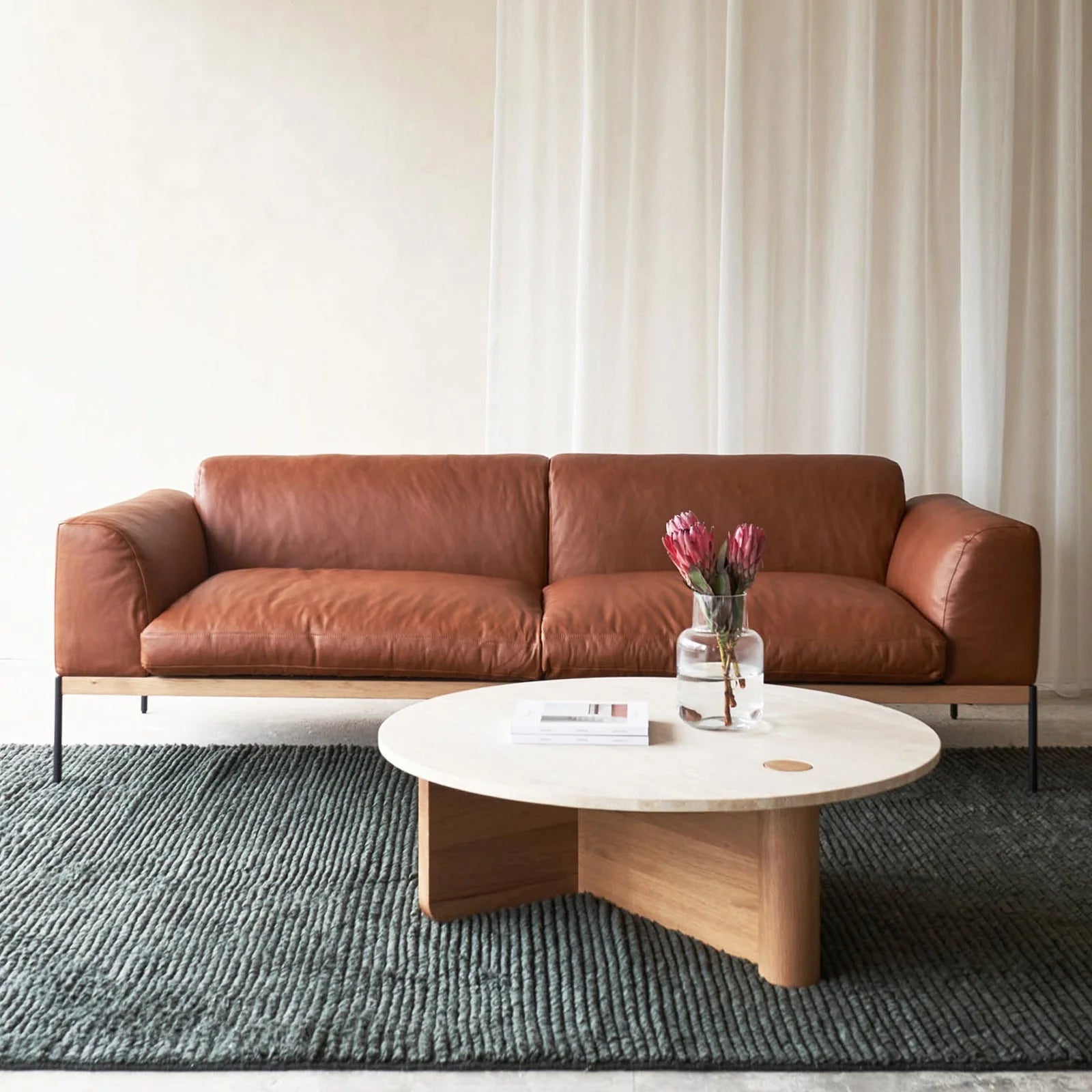 A modern living room with a Wooden Stories Department Sofa 93" in brown leather, a round white coffee table with a solid wood base, pink flowers and books on top, set on a textured gray rug with light curtains behind.
