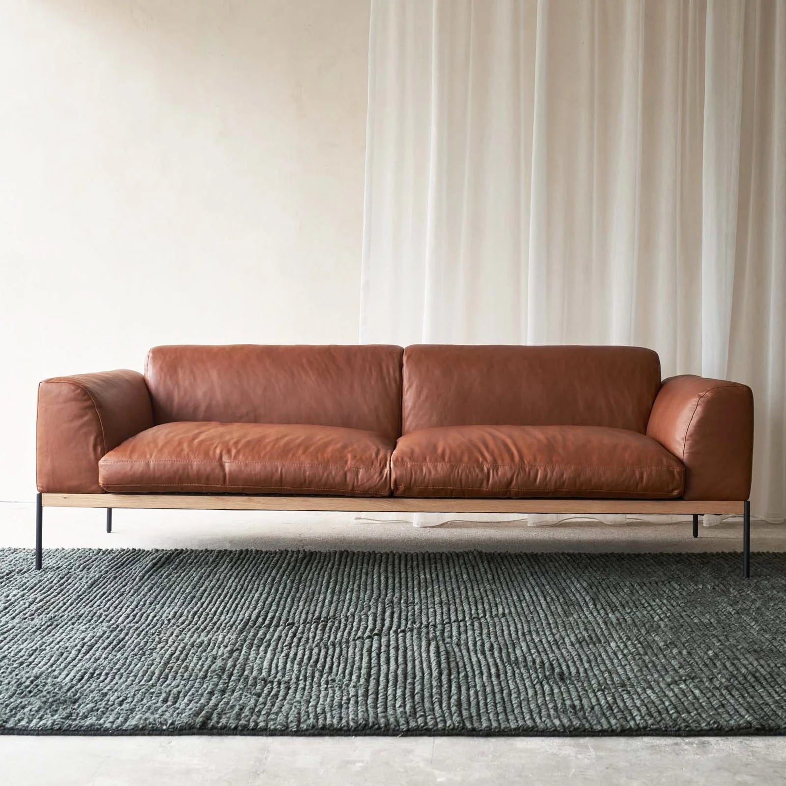 The Department Sofa 93" | Leather by Wooden Stories, featuring a modern brown leather design with a solid wood base and low black legs, sits on a textured gray rug before light walls and sheer white curtains, inspired by classic Danish style.
