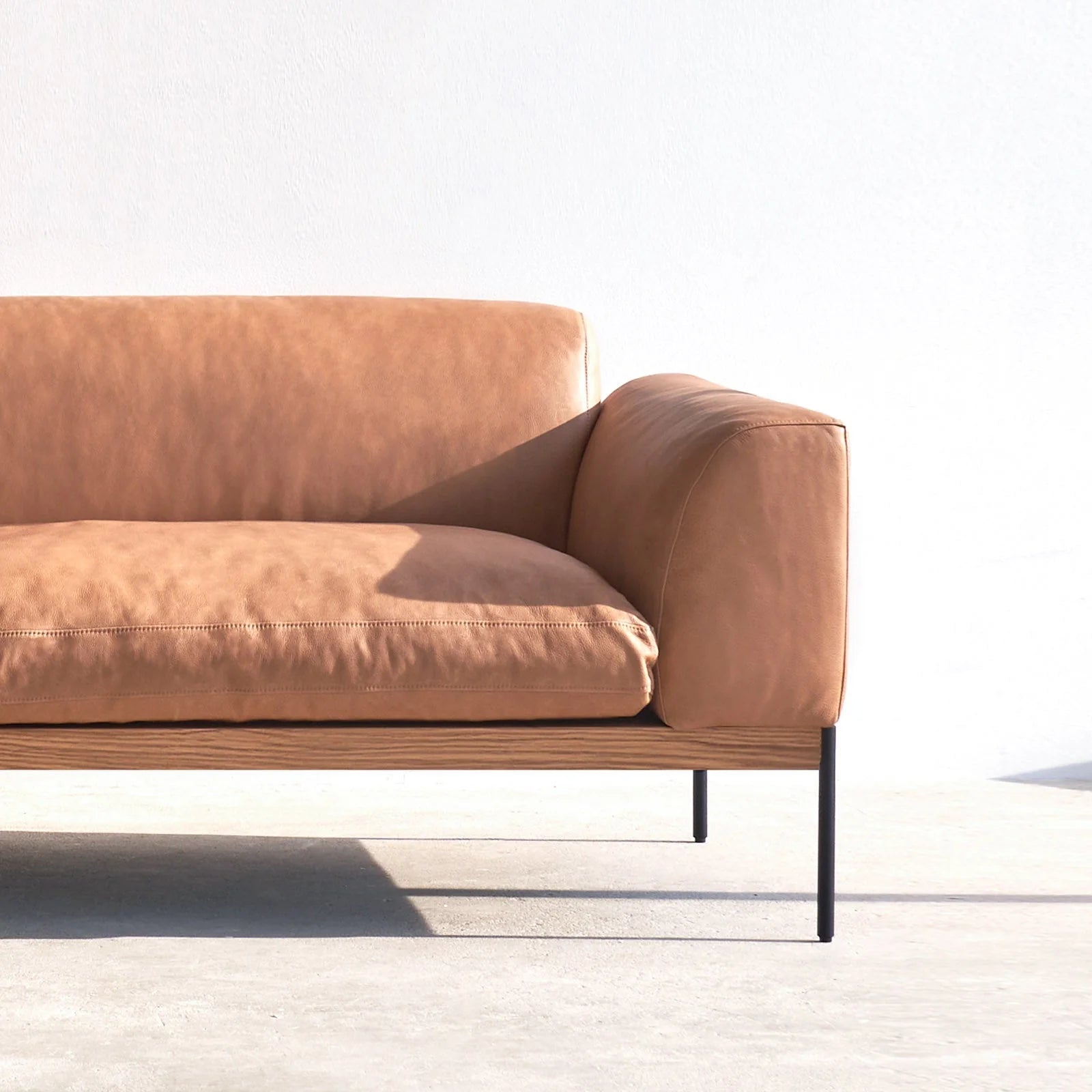 The Department Modular Sofa | Leather • M4 Left from Wooden Stories features tan leather, slim black metal legs, and a solid wood base, set against a white wall with sunlight casting floor shadows for a minimalist modern look.