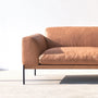 The Wooden Stories Department Modular Sofa | Leather • M5 Right features modern tan leather upholstery, slim black metal legs, and a solid wood base. Sunlight highlights its left side and seat cushion against a plain white wall on a light floor.
