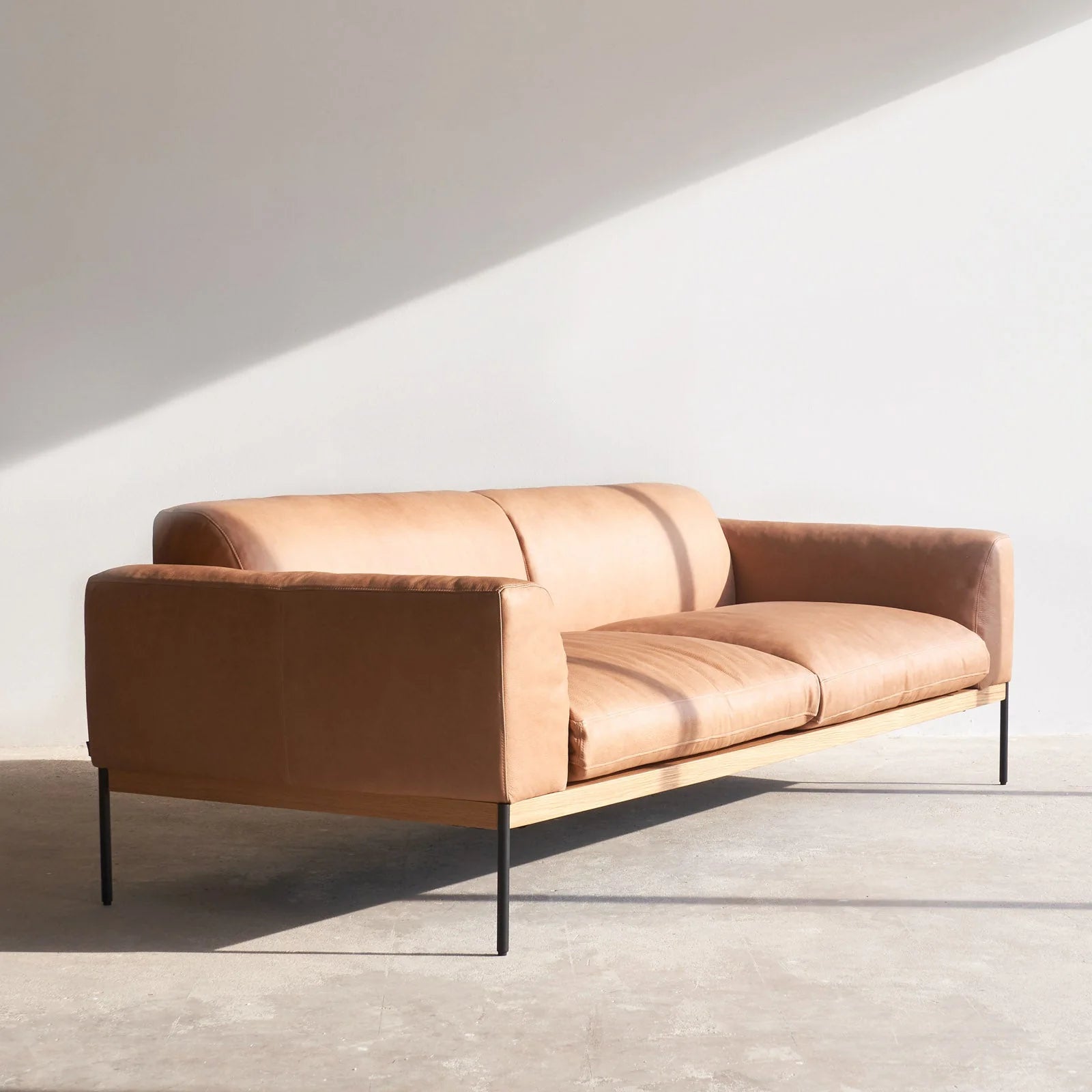 The Department Sofa 93&quot; by Wooden Stories, in tan leather with a solid wood base and black metal legs, sits in a sunlit minimal room—its sleek silhouette casting bold shadows on the white wall and gray floor.